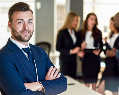 Businessman leader in modern office with businesspeople working Caucasian businessman leader looking at camera in modern office with multi-ethnic businesspeople working at the background. Teamwork concept. Young man with beard wearing blue suit.